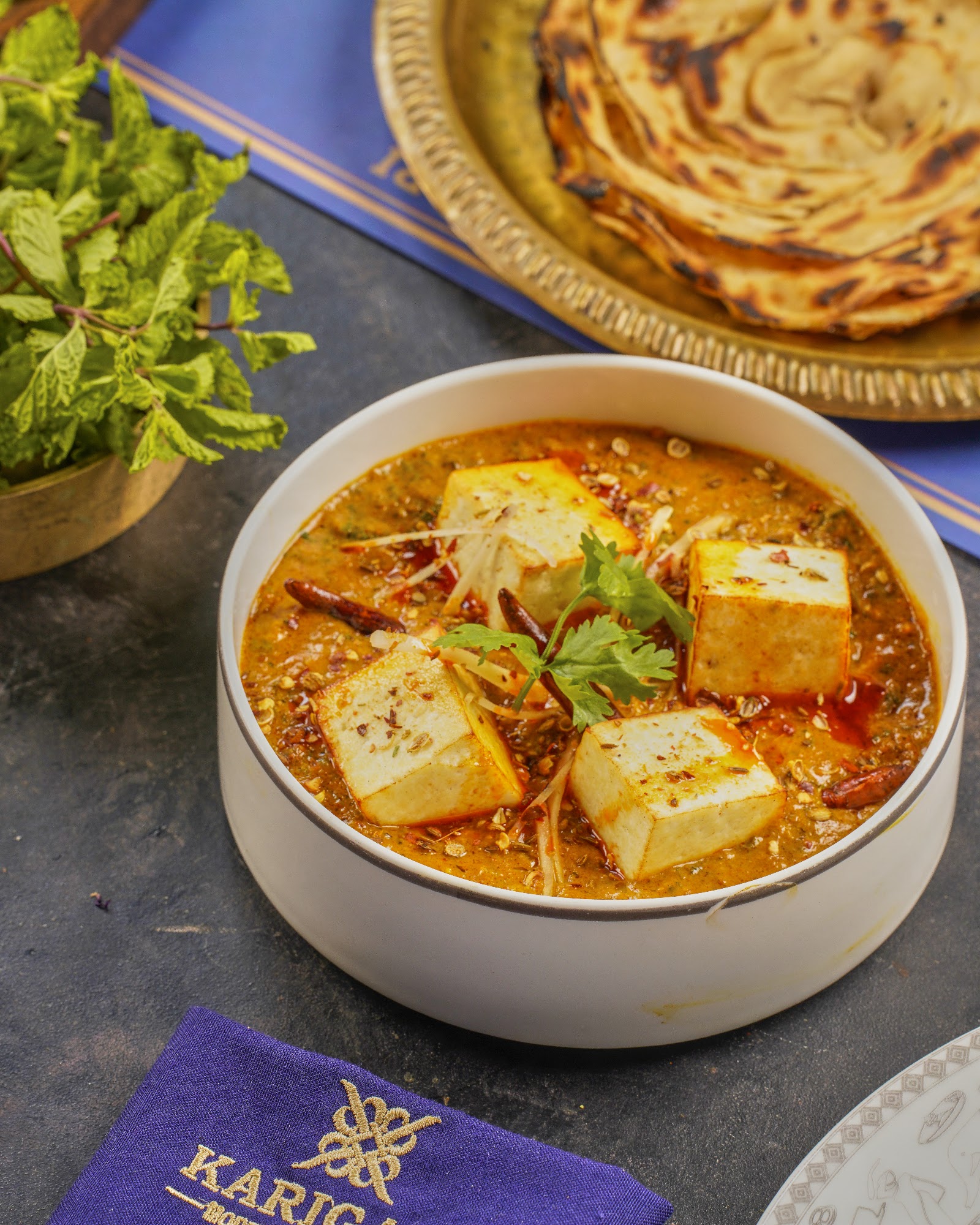 A paneer curry in a dark bowl with flatbread and fresh mint on a purple napkin